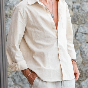 Man wearing a beige linen long sleeve casual shirt with rolled cuffs and white pants standing against stone wall